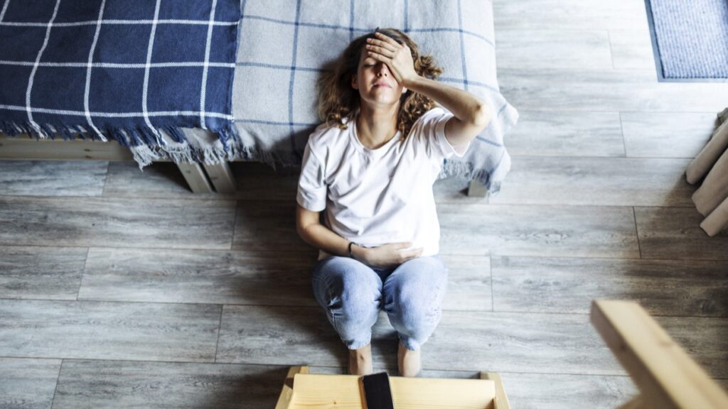 Young woman lying on the floor next to a bed, holding her head and stomach, suffering from pain due to vulvodynia